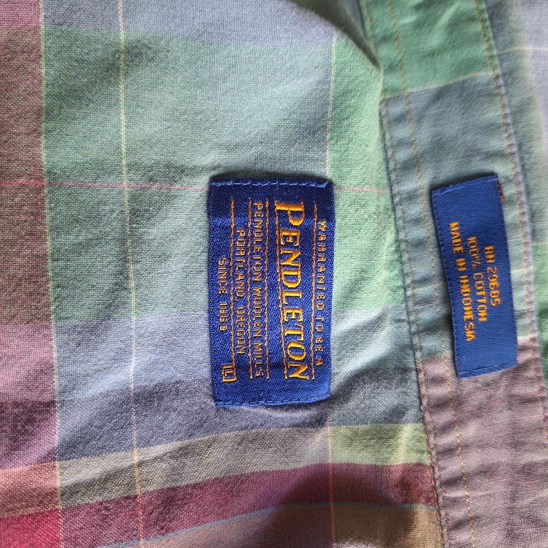 K&ouml;nnte beinhalten: Nahaufnahme eines karierten Hemdes mit einem blauen Pendleton-Etikett. Das Hemd ist eine Mischung aus Farben, darunter Gr&uuml;n, Rot und Blau. Auf dem Etikett steht "Pendleton Woolen Mills, Portland Oregon, Since 1863". Ein kleineres blaues Etikett lautet "Made in Indonesia, 100% Cotton".