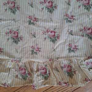 May include: A fabric textile featuring a pattern of vertical yellow and white stripes overlaid with clusters of pink roses and green leaves. The bottom edge of the fabric is finished with a gathered ruffle detail that repeats the floral pattern.