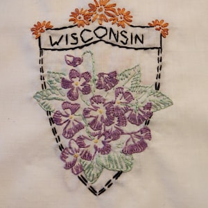 May include: Embroidered white fabric featuring a shield design with the word "WISCONSIN" in black. The shield is adorned with purple flowers, green leaves, and orange flowers at the top. The design is stitched with black thread.