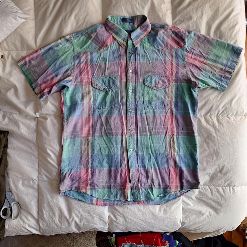 K&ouml;nnte beinhalten: Ein kurz&auml;rmeliges kariertes Hemd in Pastellfarben Rosa, Blau, Gr&uuml;n und Lila. Das Hemd hat zwei Brusttaschen, eine Knopfleiste und einen Kragen. Das Hemd ist aus einem leichten Stoff gefertigt.