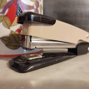 May include: A beige and black stapler with a silver metal staple holder. The stapler is on a white surface. A decorative mug with a colourful bird design is in the background.