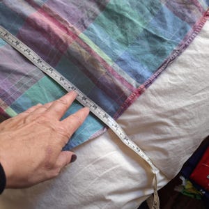 K&ouml;nnte beinhalten: Ein mehrfarbiger Stoff mit Karomuster und einem Ma&szlig;band, das eine L&auml;nge von etwa 76 cm anzeigt. Der Stoff weist ein Muster aus blauen, violetten, rosa und gr&uuml;nen Quadraten auf. Das Ma&szlig;band wird von einer Hand gehalten.