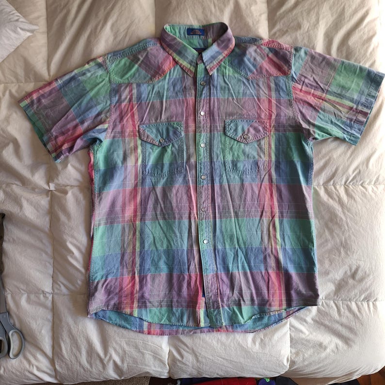 K&ouml;nnte beinhalten: Ein kurz&auml;rmeliges Hemd mit einem Karomuster in Rosa, Gr&uuml;n, Blau und Lila. Das Hemd hat zwei Brusttaschen und einen Kragen. Der Stoff scheint leicht zu sein.