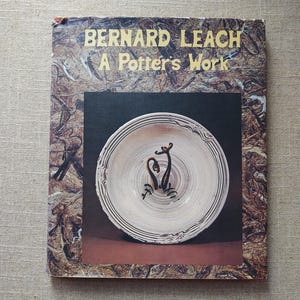 May include: A book cover featuring the title "BERNARD LEACH A Potter's Work" in gold lettering. The cover has a brown and tan textured background. A photograph of a ceramic bowl with a dark brown design is in the center.