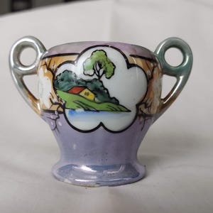 May include: A small, vintage sugar bowl with two handles. The bowl is painted with a landscape scene featuring a house, trees, and water. The exterior is a pale lavender color with silver-colored handles.