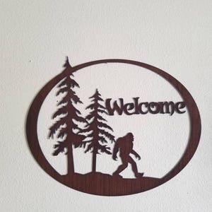 Puede incluir: Un letrero de bienvenida de metal marr&oacute;n oxidado con una silueta de Bigfoot caminando por un bosque. El letrero tiene forma de c&iacute;rculo y tiene la palabra "Welcome" escrita en &eacute;l.