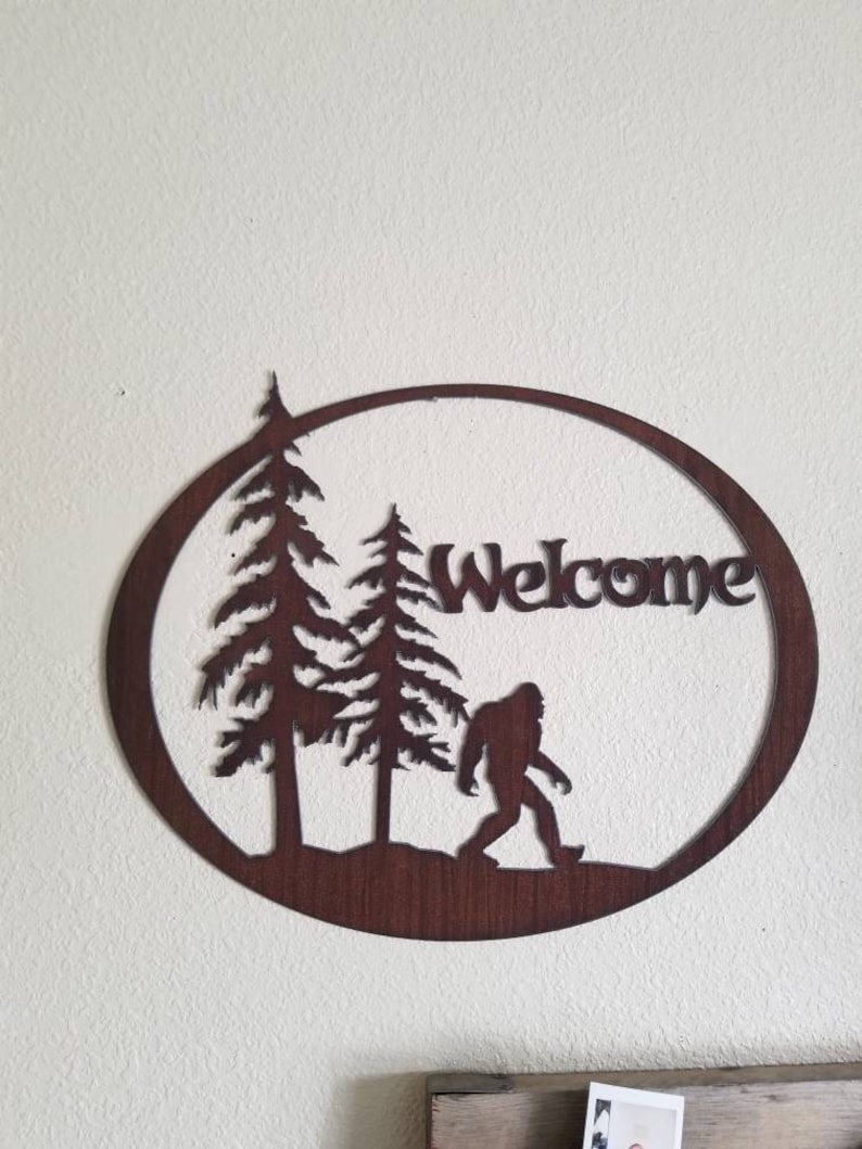 Puede incluir: Un letrero de bienvenida de metal marr&oacute;n oxidado con una silueta de Bigfoot caminando por un bosque. El letrero tiene forma de c&iacute;rculo y tiene la palabra "Welcome" escrita en &eacute;l.