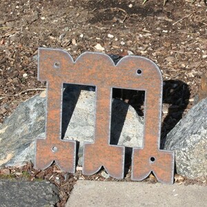 Op de afbeelding: Een verweerde, roestige metalen letter "M" met een decoratief, open ontwerp. De letter heeft een verweerde afwerking en is gemonteerd op een steen- en mulchachtergrond. Het metaal heeft een bruine en oranje kleur.