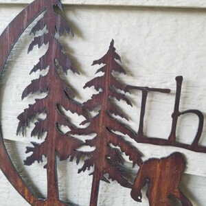 Puede incluir: Una pieza de arte de pared de metal oxidado que presenta una silueta de un Bigfoot caminando por un bosque con dos pinos. El arte tiene la forma de un c&iacute;rculo.