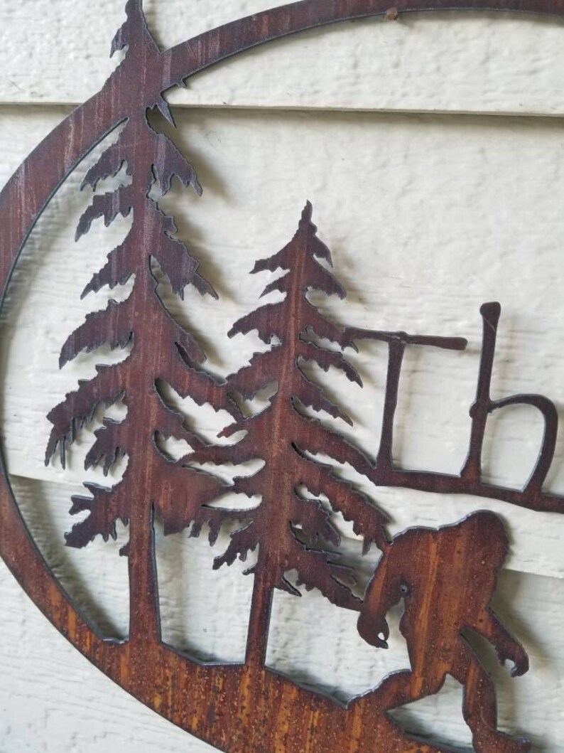 Puede incluir: Una pieza de arte de pared de metal oxidado que presenta una silueta de un Bigfoot caminando por un bosque con dos pinos. El arte tiene la forma de un c&iacute;rculo.