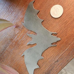 May include: A silver-colored metal bat-shaped cutout with jagged edges, resting on a wooden surface. A coin is next to the bat. The bat cutout has a detailed design, with visible wings and a head.