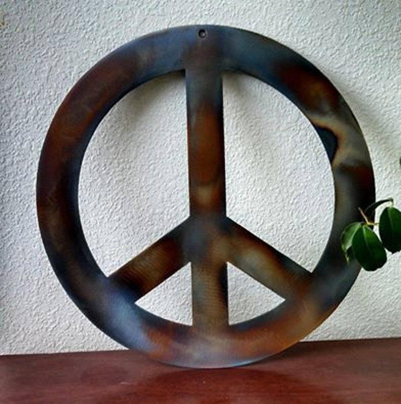 Op de afbeelding: Een groot, roestig metalen vredesteken met een blauwe en bruine patina. Het vredesteken hangt aan een witte muur.