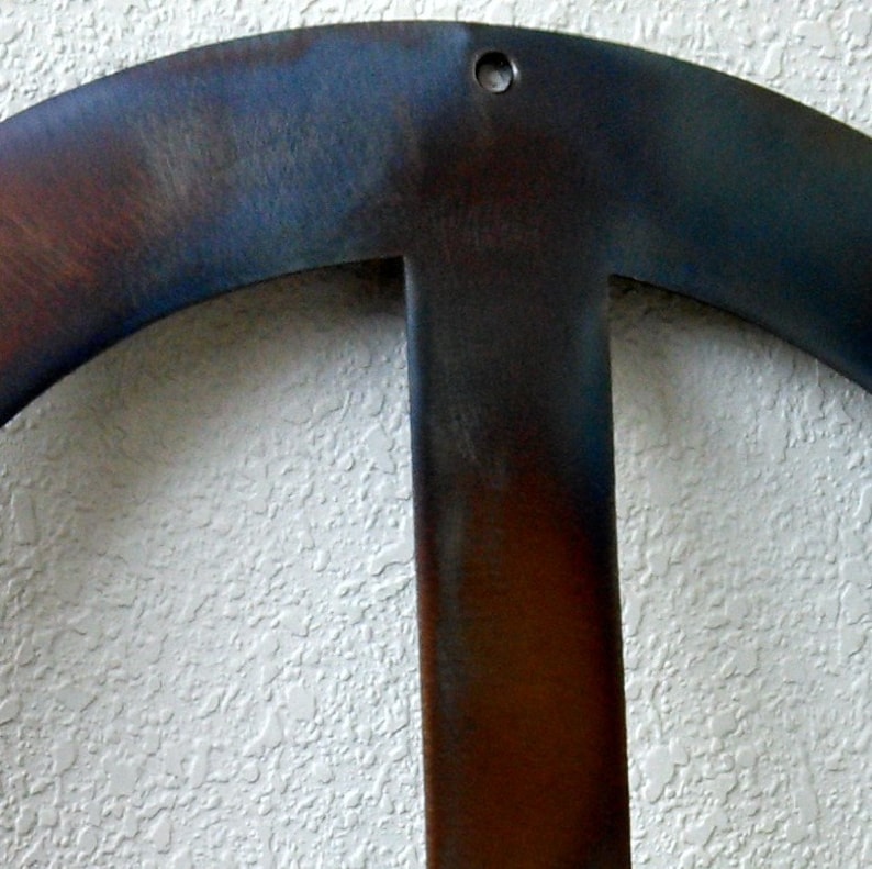Op de afbeelding: Een close-up van een metalen vredesteken met een donkerblauwe en bruine patina. Het vredesteken hangt aan een witte, getextureerde muur.