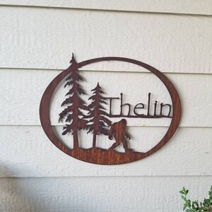 Puede incluir: Un letrero de metal marr&oacute;n oxidado con un dise&ntilde;o circular. El letrero presenta dos pinos, una silueta de Bigfoot y el nombre "Thelin" en una fuente cursiva.