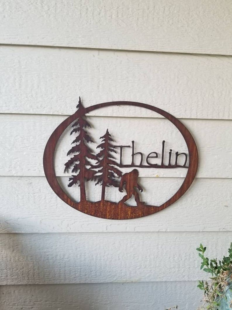 Puede incluir: Un letrero de metal marr&oacute;n oxidado con un dise&ntilde;o circular. El letrero presenta dos pinos, una silueta de Bigfoot y el nombre "Thelin" en una fuente cursiva.
