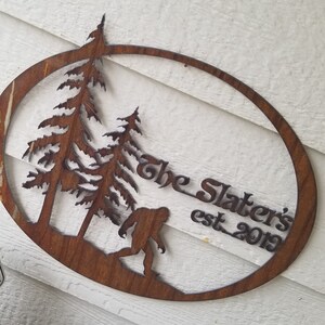 Puede incluir: Un letrero ovalado de metal marr&oacute;n oxidado con una silueta de dos pinos y un Bigfoot. El letrero dice "The Slaters est. 2019".