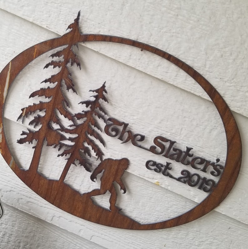 Puede incluir: Un letrero ovalado de metal marr&oacute;n oxidado con una silueta de dos pinos y un Bigfoot. El letrero dice "The Slaters est. 2019".