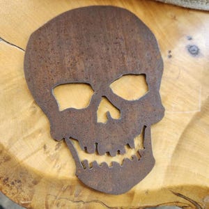 May include: A rusty brown metal skull cutout with eye sockets, a nose cavity, and a jagged jawline. The skull is placed on a light brown wooden surface, showcasing the contrast between the metal and wood.