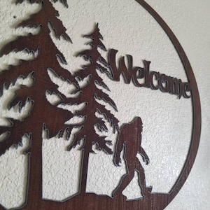 Puede incluir: Un letrero de bienvenida de metal marr&oacute;n con una silueta de Bigfoot caminando por un bosque. El letrero dice "Bienvenido" en una fuente estilizada.