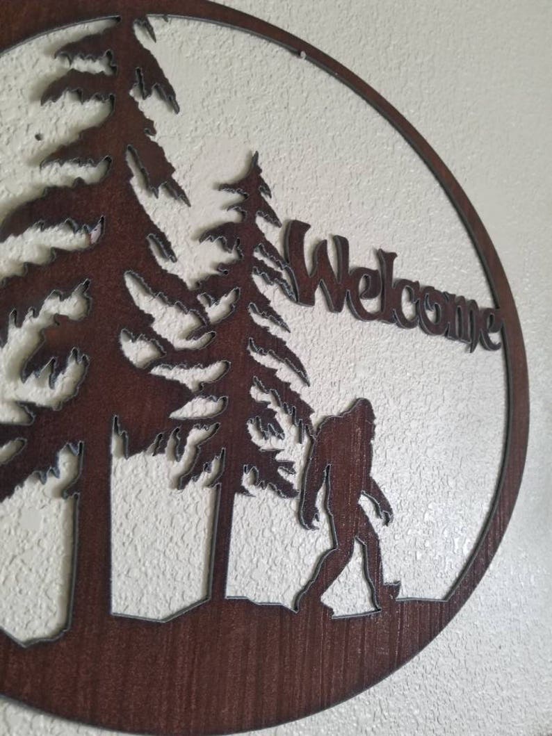 Puede incluir: Un letrero de bienvenida de metal marr&oacute;n con una silueta de Bigfoot caminando por un bosque. El letrero dice "Bienvenido" en una fuente estilizada.