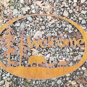 Puede incluir: Un letrero de bienvenida de metal oxidado con un dise&ntilde;o r&uacute;stico. El letrero presenta un oso, dos &aacute;rboles y tres animales m&aacute;s peque&ntilde;os. El letrero tiene forma ovalada y un fondo marr&oacute;n.