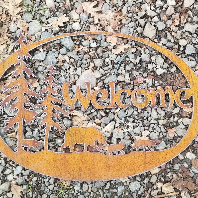 Puede incluir: Un letrero de bienvenida de metal oxidado con un dise&ntilde;o r&uacute;stico. El letrero presenta un oso, dos &aacute;rboles y tres animales m&aacute;s peque&ntilde;os. El letrero tiene forma ovalada y un fondo marr&oacute;n.