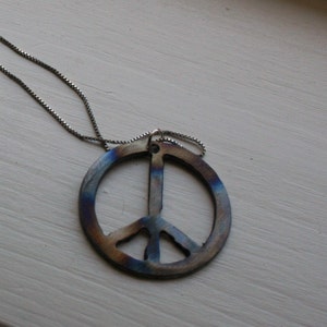Op de afbeelding: Een zilveren ketting met een vredesteken hanger. De hanger is gemaakt van metaal en heeft een blauwe, paarse en bruine iriserende afwerking.