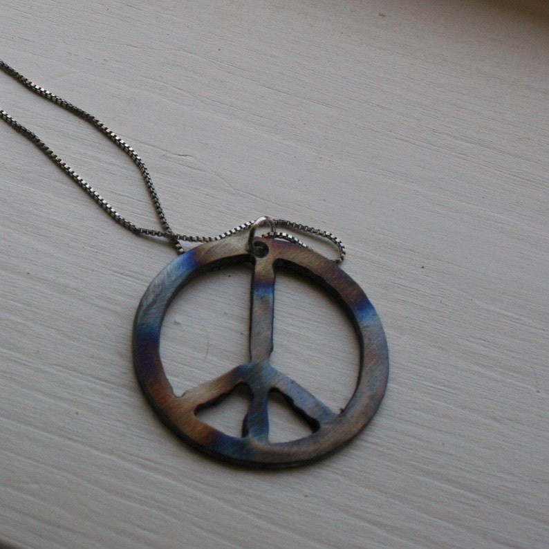 Op de afbeelding: Een zilveren ketting met een vredesteken hanger. De hanger is gemaakt van metaal en heeft een blauwe, paarse en bruine iriserende afwerking.