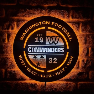 May include: Circular illuminated sign with the text "WASHINGTON FOOTBALL" and "COMMANDERS". The sign displays the team's logo, the year "1932", and the years 1937, 1942, 1952, and 1991. It is lit with an orange glow against a brick wall.