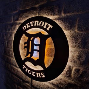 Puede incluir: Logotipo de los Detroit Tigers en blanco y negro con una 'D' iluminada en el centro. El logotipo est&aacute; en una se&ntilde;al redonda de madera negra.