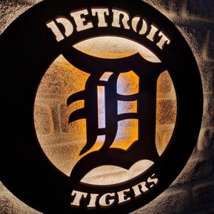 Puede incluir: Un letrero circular blanco y negro con las palabras "DETROIT TIGERS" en letras blancas. El centro del letrero presenta una gran "D" blanca con un contorno negro. El letrero est&aacute; iluminado desde atr&aacute;s, creando un brillo c&aacute;lido.