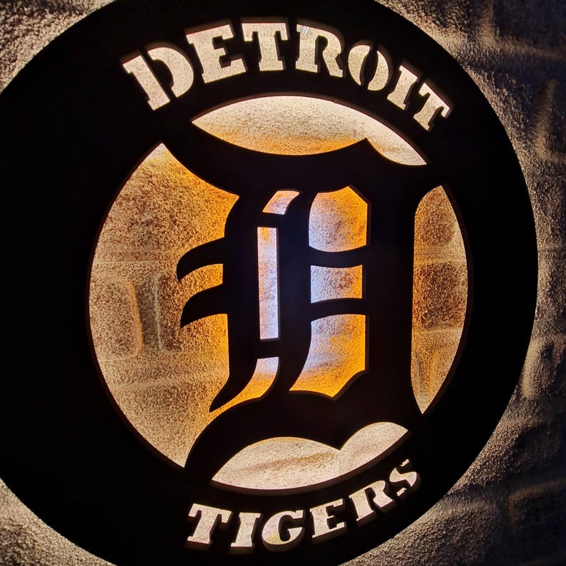 Puede incluir: Un letrero circular blanco y negro con las palabras "DETROIT TIGERS" en letras blancas. El centro del letrero presenta una gran "D" blanca con un contorno negro. El letrero est&aacute; iluminado desde atr&aacute;s, creando un brillo c&aacute;lido.