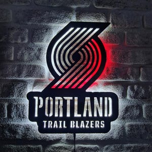 Puede incluir: Un letrero iluminado en blanco y negro con el logotipo de los Portland Trail Blazers y el nombre del equipo. El logotipo es un diseño estilizado con detalles en rojo, y el texto es en negrita e iluminado. Montado en una pared de ladrillo.