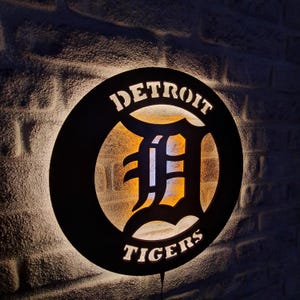 Puede incluir: Un letrero circular negro con un logotipo de Detroit Tigers blanco. El logotipo presenta una gran "D" blanca con un contorno negro y las palabras "Detroit" y "Tigers" en texto blanco.