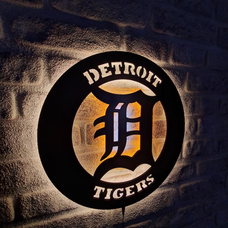 Puede incluir: Un letrero circular negro con un logotipo de Detroit Tigers blanco. El logotipo presenta una gran "D" blanca con un contorno negro y las palabras "Detroit" y "Tigers" en texto blanco.