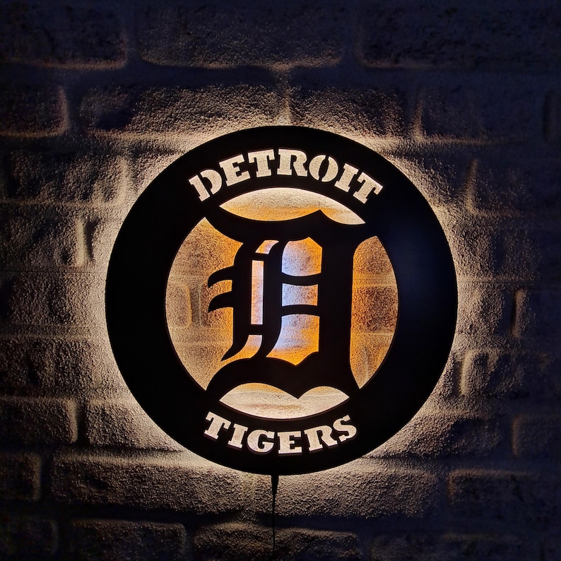 Puede incluir: Un letrero circular negro con un contorno blanco y las palabras "DETROIT TIGERS" en una fuente estilizada. El centro del letrero presenta una "D" blanca con un contorno negro. El letrero est&aacute; iluminado desde atr&aacute;s, creando un brillo c&aacute;lido.