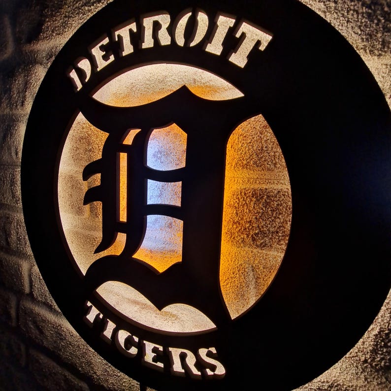 Puede incluir: Un letrero de metal negro con las palabras "DETROIT TIGERS" en letras blancas. El letrero presenta una gran "D" estilizada en el centro, con una luz que brilla a trav&eacute;s de la forma recortada.