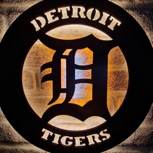 Puede incluir: Un letrero circular negro con las palabras "DETROIT TIGERS" en letras blancas. El centro del letrero presenta una gran "D" estilizada negra con un brillo amarillo detr&aacute;s.