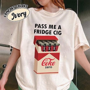 May include: Ivory-colored t-shirt with the text "PASS ME A FRIDGE CIG". The shirt features a graphic of a red and beige cigarette box filled with Coke Zero cans. The words "Coke zero" are printed on the box.