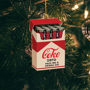 May include: A novelty Christmas ornament shaped like a pack of cigarettes, with a red and white design. The pack is open, revealing miniature Coke Zero cans. The text "Coke Zero" and "PASS ME A FRIDGE CIG" are printed on the front.