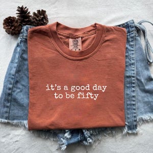 Peut inclure: Un t-shirt à manches courtes de couleur rouille avec le texte blanc "it's a good day to be fifty" imprimé sur le devant. Le t-shirt présente un col rond et une étiquette de marque Comfort Colors à l'intérieur, posé sur une jupe en jean bleu.
