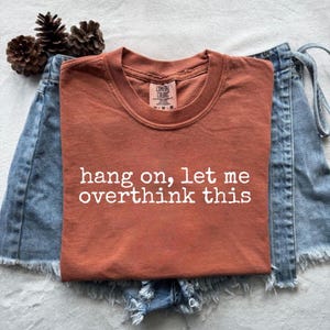 Puede incluir: Camiseta Comfort Colors de color óxido con la frase "hang on, let me overthink this" en letras blancas. La camiseta se combina con pantalones cortos vaqueros y piñas.