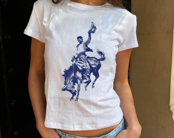 May include: White t-shirt with a navy blue illustration of a rodeo cowboy on a bucking bronco. The cowboy is holding his hat aloft. The top has a crew neck and short sleeves.