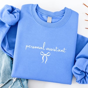 May include: A periwinkle blue crewneck sweatshirt featuring the white text personal assistant in a cursive script font, positioned above a simple white line-drawn bow graphic centered on the chest of the garment.