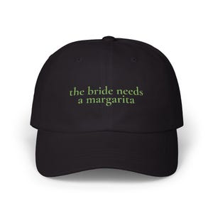 Może przedstawiać: Czarna czapka z daszkiem z wyhaftowanym napisem "the bride needs a margarita" jasnozieloną czcionką, umieszczonym centralnie na przednim panelu. Czapka posiada zakrzywiony daszek, usztywnianą koronę oraz guzik na samym szczycie główki.