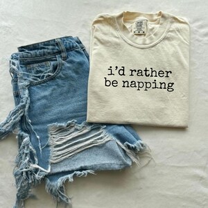 Pode incluir: Um conjunto casual com calções de ganga azul claro com detalhes desgastados e uma t-shirt creme. A t-shirt tem a frase "i'd rather be napping" impressa em preto, perfeita para um look descontraído e diário.