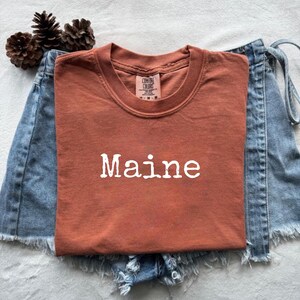 Może przedstawiać: Rdzawa koszulka z białym napisem "Maine". Koszulka jest prezentowana na dżinsowych szortach. Obraz zawiera również szyszki. Koszulka jest marki Comfort Colors.
