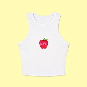 Pode incluir: Top curto branco com um gráfico de maçã vermelha. A maçã tem uma folha verde e as letras "NYC" em rosa. O top sem mangas tem decote redondo.