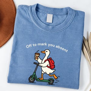 May include: A blue crew neck t-shirt featuring a graphic of a white duck with an orange beak and feet, wearing a red backpack, holding a brown coffee cup, and riding a green scooter. The text above the duck reads, Off to mark you absent.
