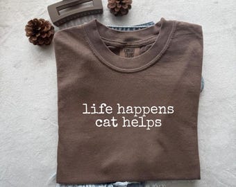 Puede incluir: Una camiseta marrón con el texto "life happens cat helps" en blanco. La camiseta está doblada sobre un pantalón corto vaquero azul. En la imagen también hay una pinza para el pelo marrón y dos piñas pequeñas.